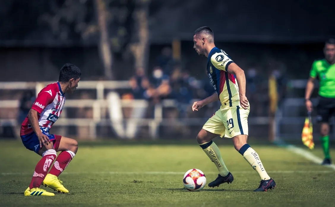Adrian Goransch, número 29, con la camiseta crema y azul marino del Club América, frente a un rival con rayas rojas y blancas