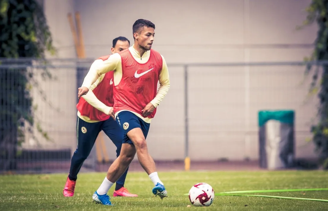 Adrian Goransch golpeando un balón con un peto de entrenamiento rojo del Club América, con un compañero detrás