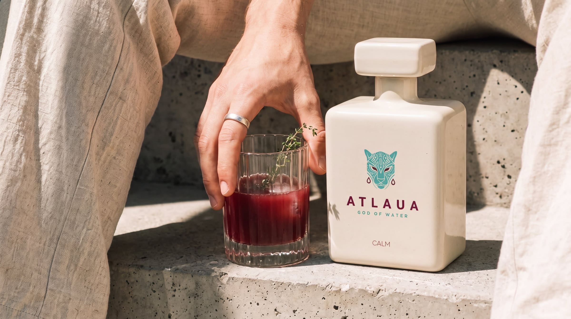 Una mano levantando un vaso de ATLAUA junto a la botella de CALM sobre escalones de piedra bañados de sol