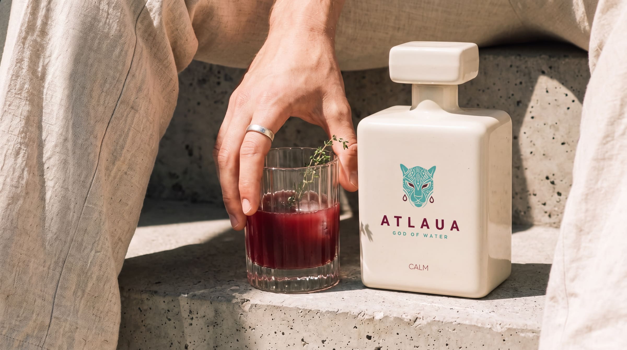 Una mano apoyada en un vaso estriado de jugo de frutos rojos, junto a una botella de ATLAUA CALM sobre escalones de piedra bañados de sol