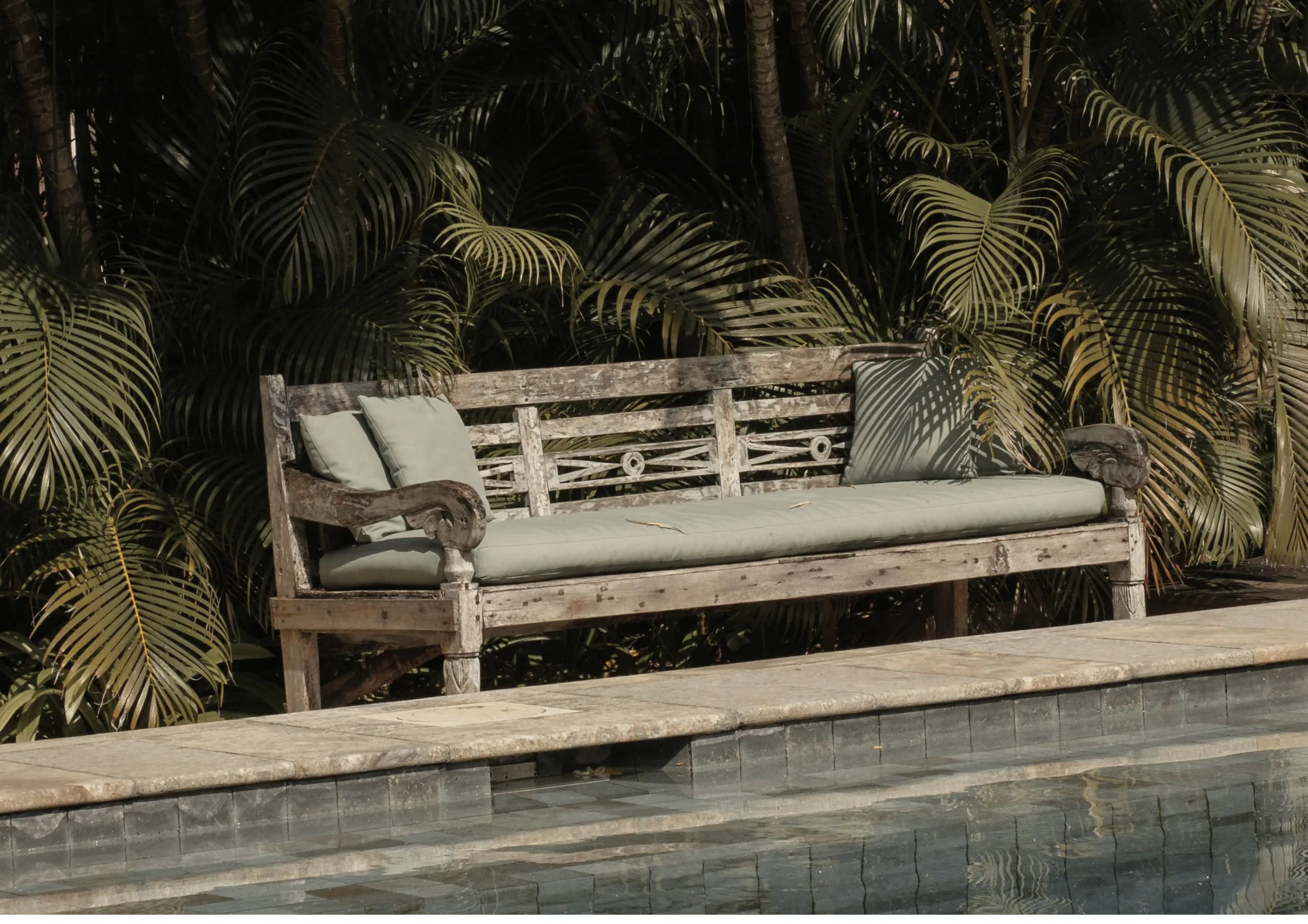 Un banco de teca desgastado con cojines color salvia junto a una piscina, con palmeras detrás bajo el sol de la tarde